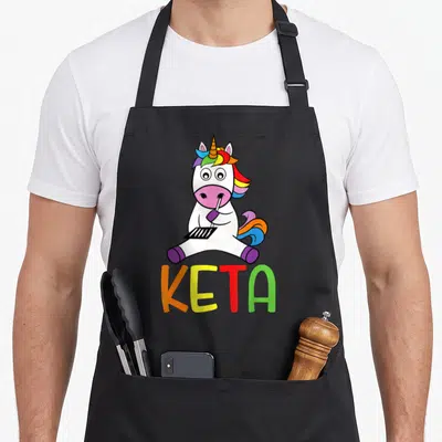 Tablier de cuisine noir avec une licorne blanche buvant dans une paille au-dessus du mot KETA multicolore.