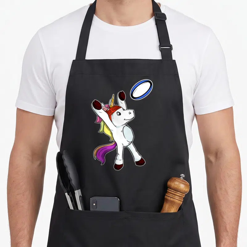 Tablier de cuisine noir avec une licorne blanche portant un casque rouge et attrapant un ballon de rugby ovale bleu et blanc.