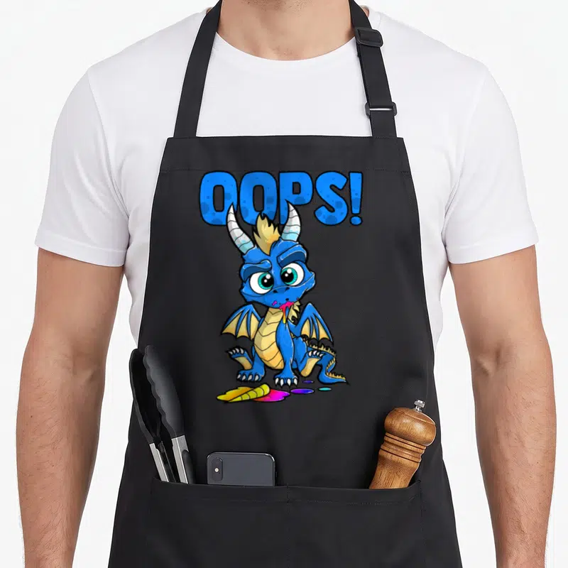 Tablier de cuisine noir avec un dragon bleu mangeant une licorne colorée et le texte Oups en lettres capitales.