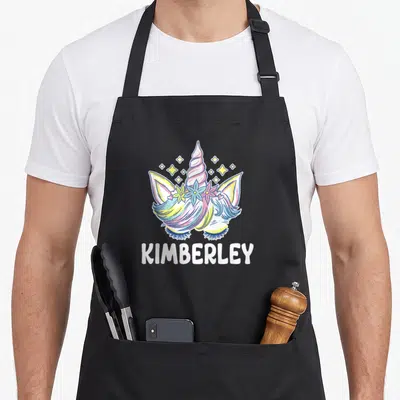 Tablier de cuisine noir avec une licorne aux cheveux multicolores, une couronne de fleurs et le nom Kimberley en lettres blanches en dessous.