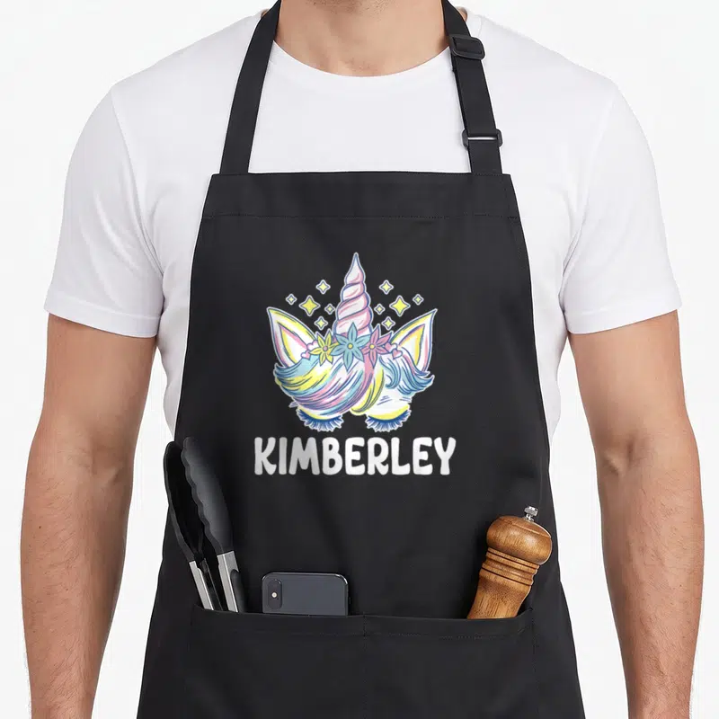 Tablier de cuisine noir avec une licorne aux cheveux multicolores, une couronne de fleurs et le nom Kimberley en lettres blanches en dessous.