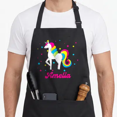 Tablier de cuisine noir avec licorne arc-en-ciel rose et bleue et le prénom Amelia en lettres roses entouré d'étoiles.