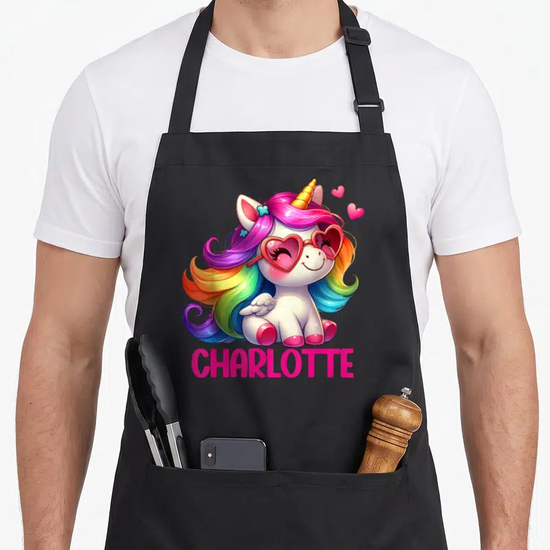 Tablier de cuisine noir avec une licorne aux cheveux multicolores, lunettes cœurs et le nom Charlotte en lettres roses vives.