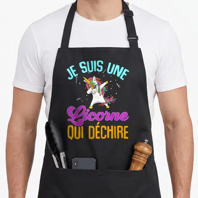 Tablier de cuisine noir avec une licorne blanche effectuant un dab et du texte multicolore arqué au-dessus et en dessous de l'illustration.