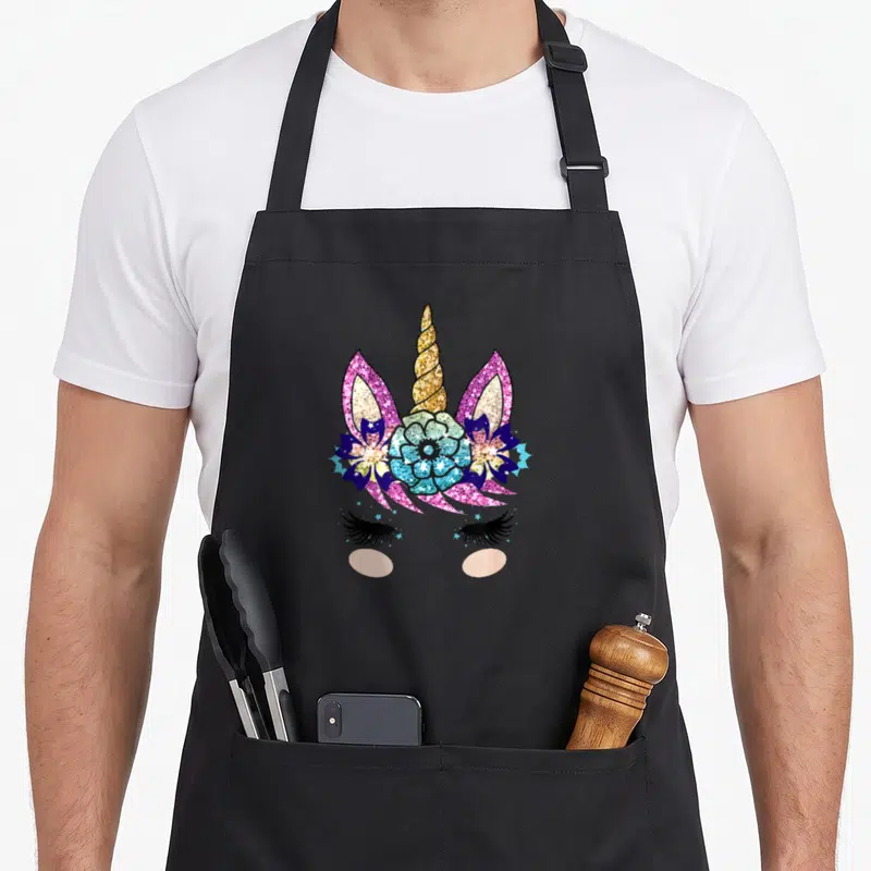 Tablier de cuisine noir avec visage de licorne aux cils, corne dorée et fleurs pailletées roses et bleues.