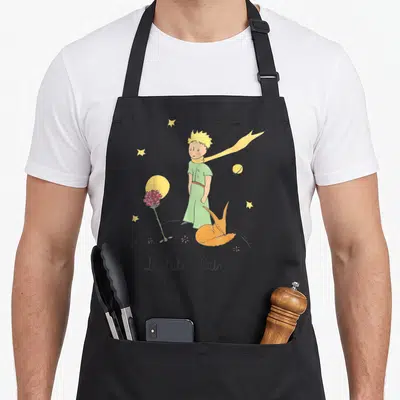 Tablier de cuisine noir avec illustration du Petit Prince, du renard orange, d'une rose rouge et des étoiles jaunes sur fond gris.
