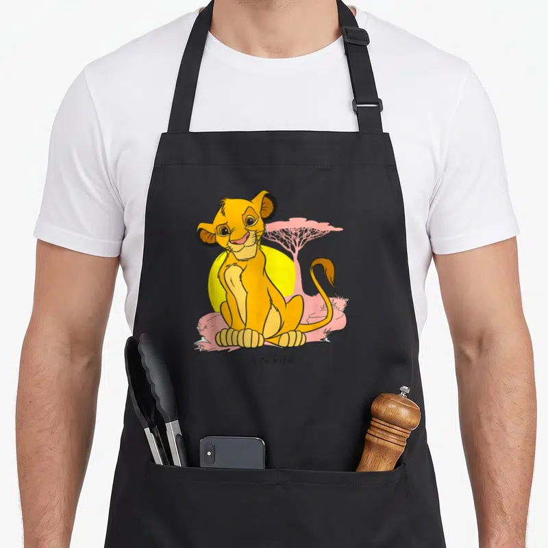 Tablier de cuisine noir avec un jeune lion orange assis devant un soleil jaune et un arbre rose pastel.