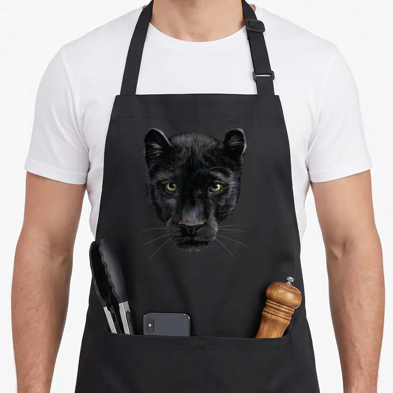 Tablier de cuisine noir avec tête de panthère noire réaliste aux yeux verts et poils détaillés.