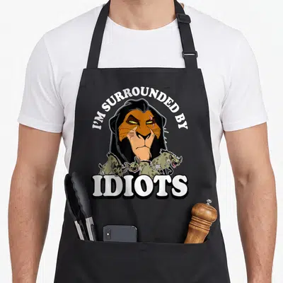 Tablier de cuisine noir avec le personnage Scar entouré d'hyènes et le texte I'M SURROUNDED BY IDIOTS imprimé en blanc.