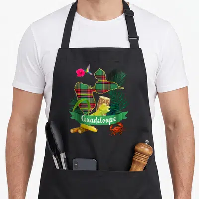 Tablier de cuisine noir avec une carte de Guadeloupe en motif madras, un colibri, des feuilles de palmier et un crabe rouge.