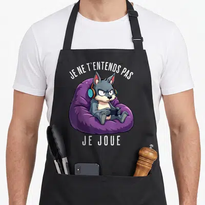 Tablier de cuisine noir avec un loup gris assis sur un coussin violet portant des écouteurs et tenant une manette.