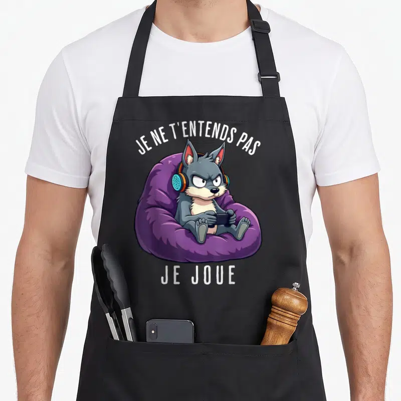 Tablier de cuisine noir avec un loup gris assis sur un coussin violet portant des écouteurs et tenant une manette.