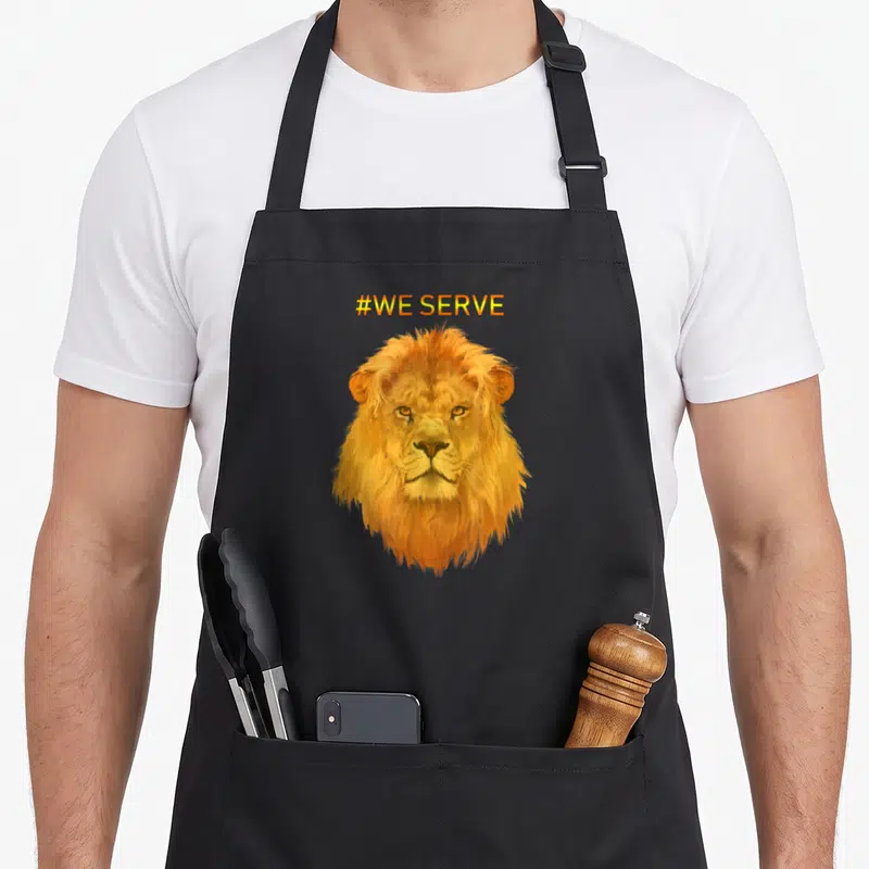 Tablier de cuisine noir avec tête de lion orange et texte #WE SERVE en haut.