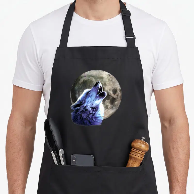 Tablier de cuisine noir avec un loup hurlant aux teintes bleues devant une lune texturée.