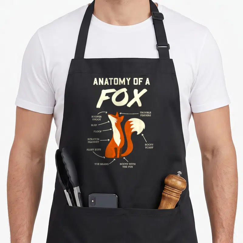 Tablier de cuisine noir avec une illustration d'un renard orange et blanc accompagné de légendes humoristiques en anglais.