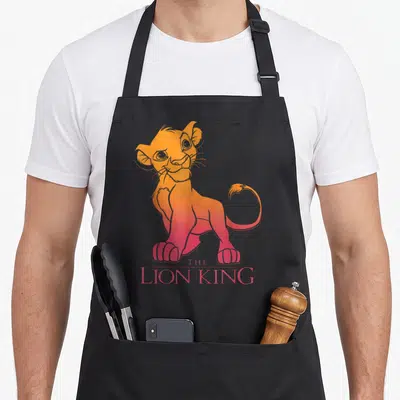 Tablier de cuisine noir avec un portrait de Simba en dégradé orange et rose sous le titre The Lion King.