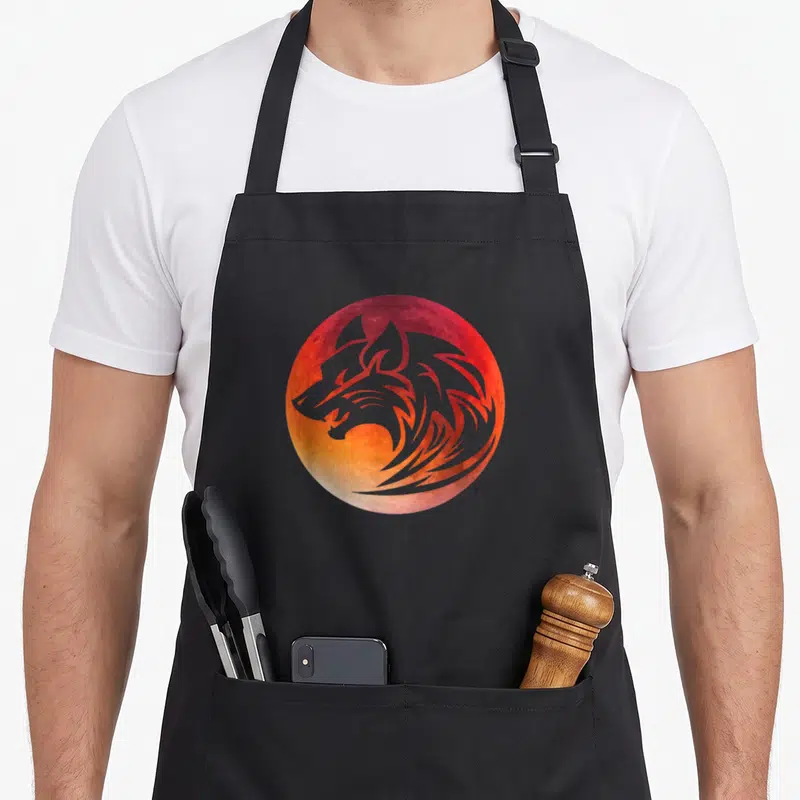 Tablier de cuisine noir avec illustration circulaire d'un loup tribal gris devant une lune rouge et orange texturée.