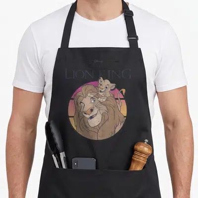 Tablier de cuisine noir avec illustration circulaire de Mufasa et Simba sur fond dégradé rose et orange.