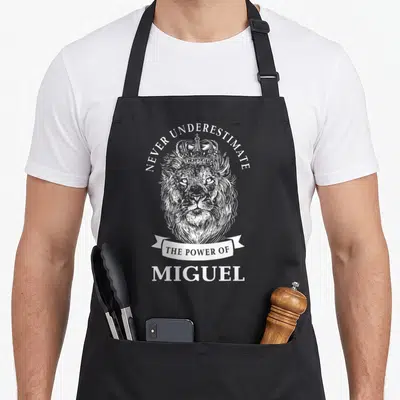 Tablier de cuisine noir avec un lion couronné et le texte Never Underestimate The Power Of Miguel imprimé en blanc.