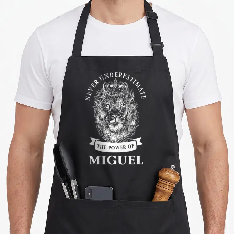 Tablier de cuisine noir avec un lion couronné et le texte Never Underestimate The Power Of Miguel imprimé en blanc.