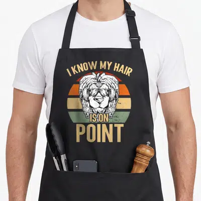 Tablier de cuisine noir avec tête de lion aux dreadlocks, lunettes de soleil et texte I KNOW MY HAIR IS ON POINT sur fond de disque solaire retro.