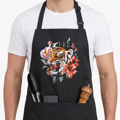 Tablier de cuisine noir avec illustration d'un tigre aux yeux jaunes entouré de fleurs roses et de feuilles bleutées.