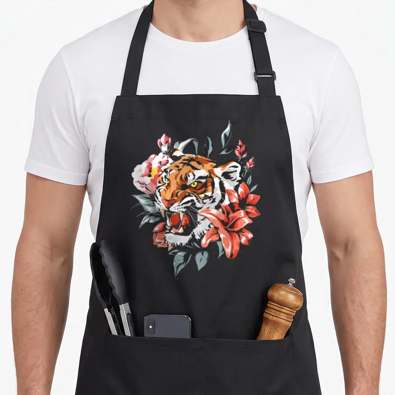 Tablier de cuisine noir avec illustration d'un tigre aux yeux jaunes entouré de fleurs roses et de feuilles bleutées.