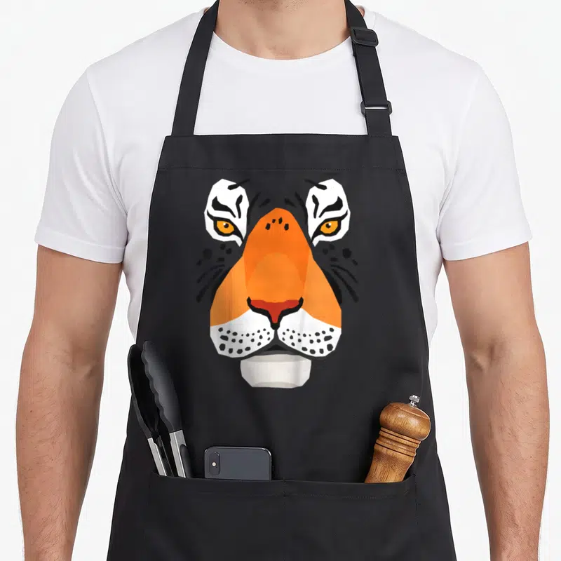 Tablier de cuisine noir avec un visage de tigre géométrique orange, blanc et noir aux yeux perçants.