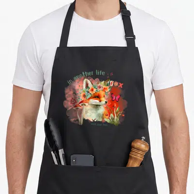 Tablier de cuisine noir avec illustration de renard floral, champignons rouges et papillon rose sur fond nuageux.