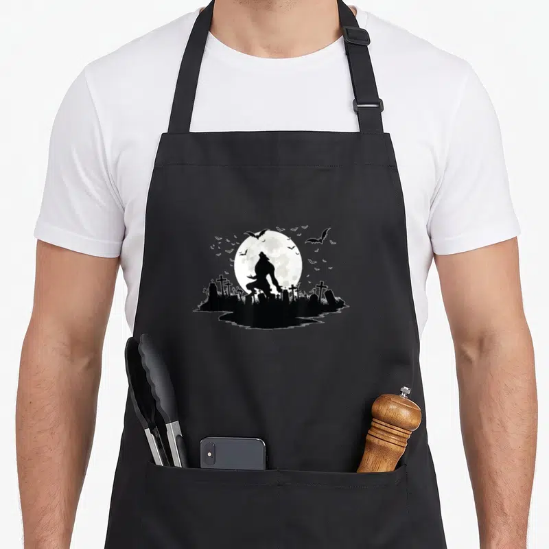 Tablier de cuisine noir avec silhouette de loup-garou sous une pleine lune au-dessus d'un cimetière.