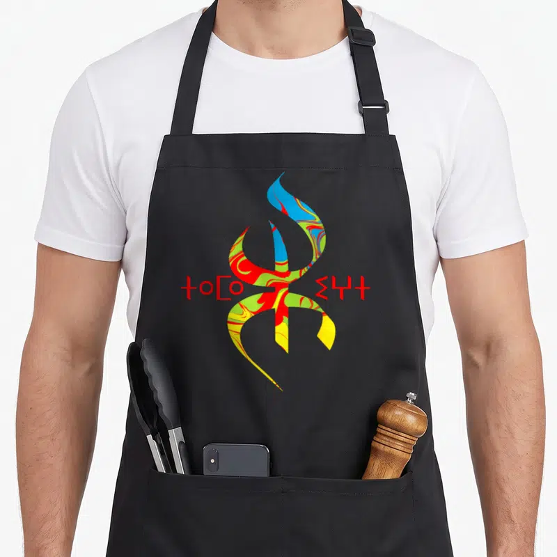 Tablier de cuisine noir avec un grand symbole Tifinagh multicolore et des inscriptions en caractères rouges sur le côté.