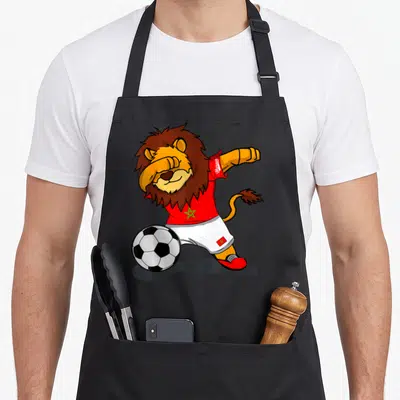 Tablier de cuisine noir avec un lion footballeur marocain en tenue rouge et verte dribblant un ballon.