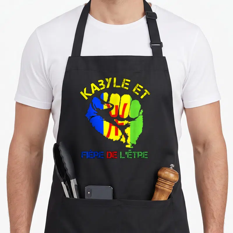 Tablier de cuisine noir avec un poing aux couleurs kabyles et le texte Ka3yle et fière de l'être imprimé en jaune et multicolore.