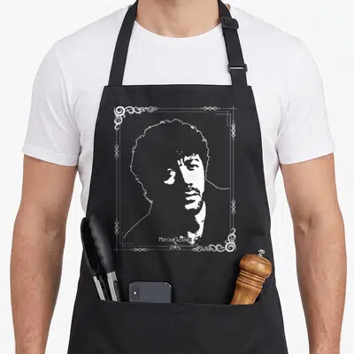 Tablier de cuisine noir avec portrait blanc de Matoub Lounes encadré de motifs ornementaux traditionnels berbères sur fond uni.