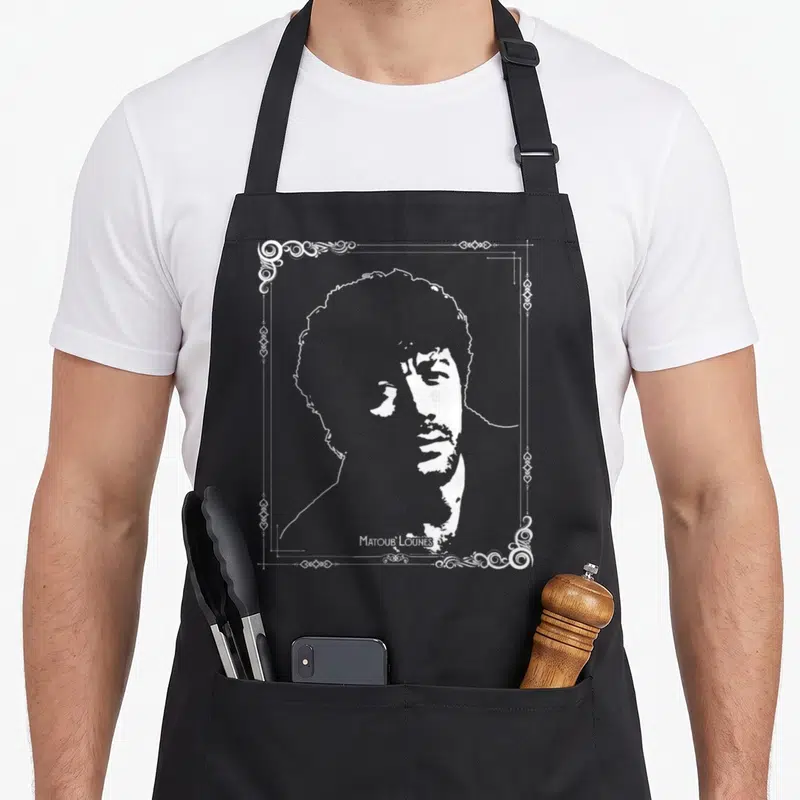 Tablier de cuisine noir avec portrait blanc de Matoub Lounes encadré de motifs ornementaux traditionnels berbères sur fond uni.