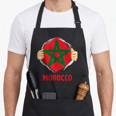 Tablier de cuisine noir avec illustration de mains déchirant le tissu pour révéler un drapeau marocain rouge et vert surmonté du mot Morocco.