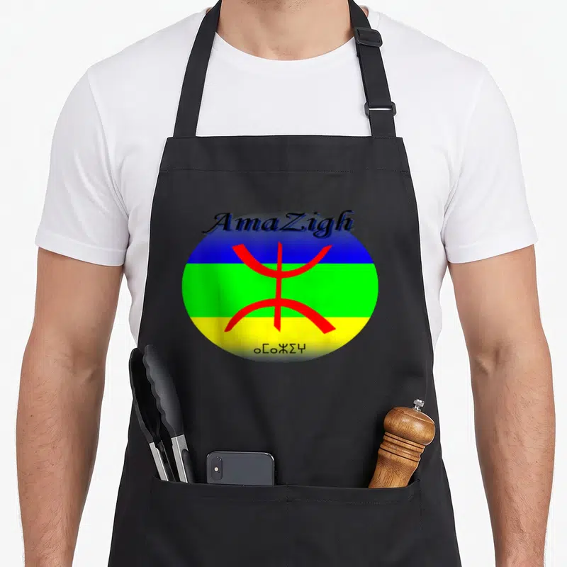 Tablier de cuisine noir avec le drapeau amazigh circulaire, le signe Tifinagh rouge et les mots AmaZigh et Tamazgha imprimés.