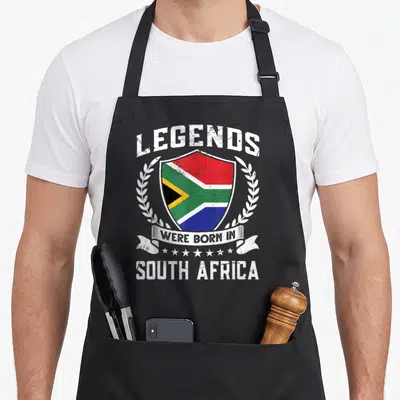 Tablier de cuisine noir avec un écu coloré portant le drapeau sud-africain et des lettres blanches vintage.