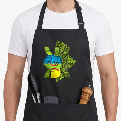 Tablier de cuisine noir avec un poing coloré aux teintes kabyles superposé à des ornements tifinagh verts sur fond uni.