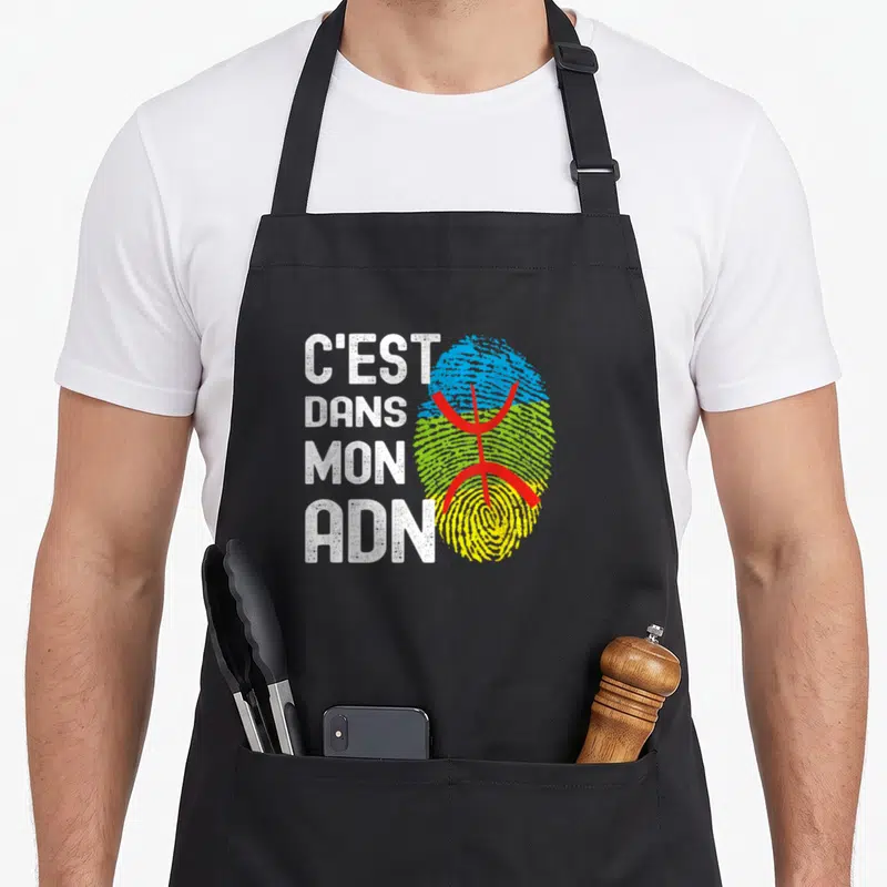 Tablier de cuisine noir avec l'inscription C'est Dans Mon ADN et un drapeau berbère stylisé en empreinte digitale.
