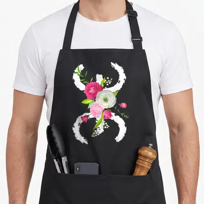 Tablier de cuisine noir avec le symbole floral berbère azigh kabyle entouré de roses roses et blanches ainsi que de feuilles vertes.