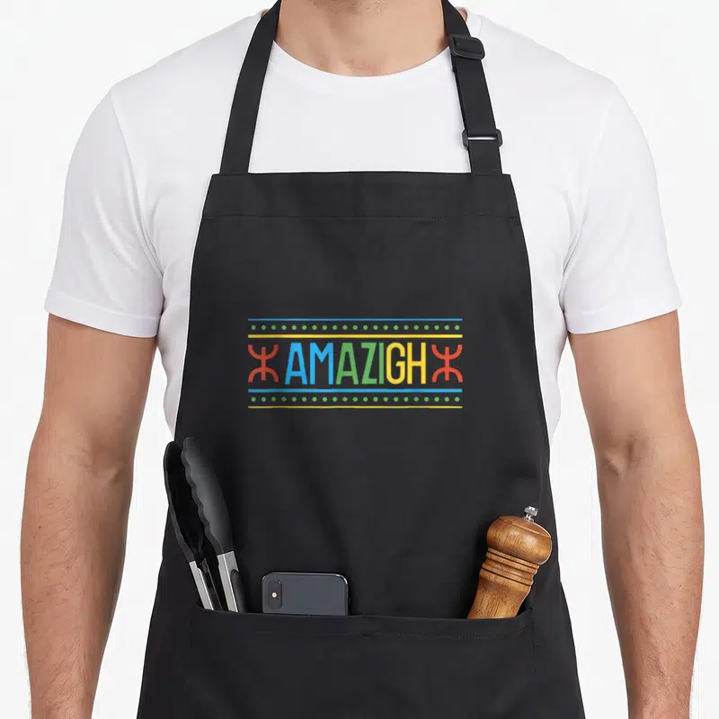 Tablier de cuisine noir avec inscription AMAZIGH multicolore et symboles tifinagh rouges sur fond gris uni.