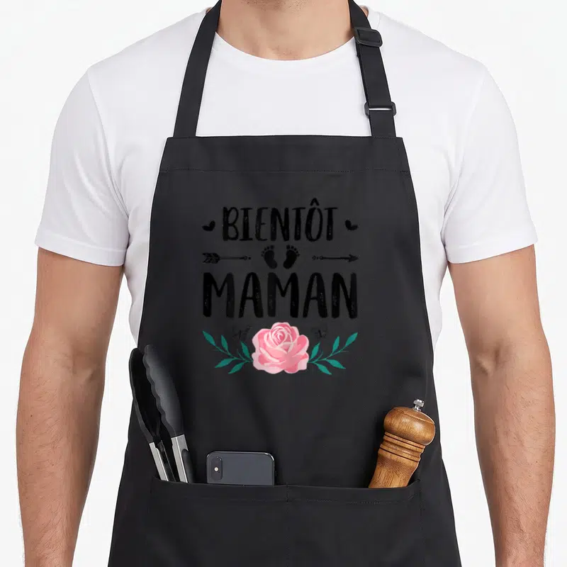 Tablier de cuisine noir avec l'inscription Bientôt Maman, une rose rose, des papillons et des empreintes de pieds en bas du motif.