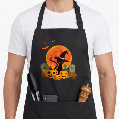 Tablier de cuisine noir avec illustration Halloween montrant un chat sorcière, des citrouilles et des tombes devant une lune orange.