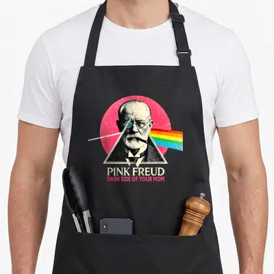 Tablier de cuisine noir avec portrait de Freud dans un triangle émettant un arc-en-ciel sur fond rose.