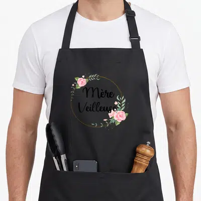 Tablier de cuisine noir avec inscription Mère Veilleuse entourée de roses roses et de feuillage vert dans un cercle doré.