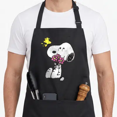 Tablier de cuisine noir avec Snoopy tenant des fleurs roses et Woodstock en vol au-dessus.