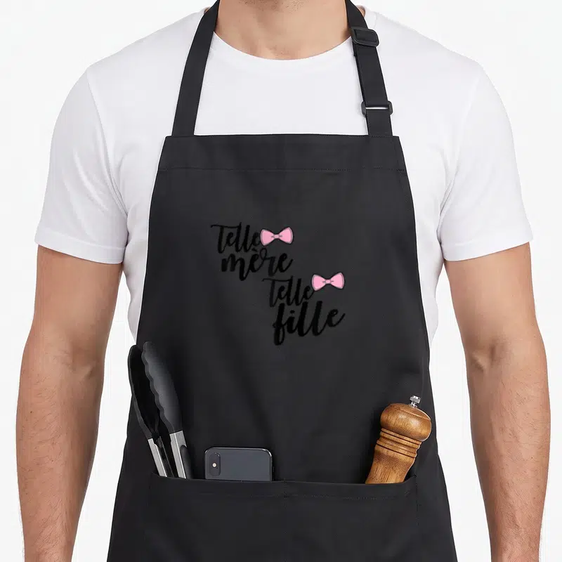Tablier de cuisine noir avec inscription Telle mère telle fille et deux nœuds roses stylisés.