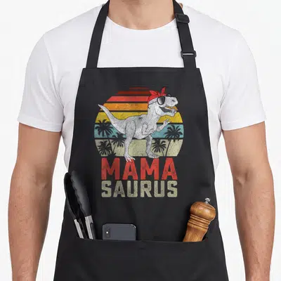 Tablier de cuisine noir avec un T-Rex portant un bandeau rouge, des lunettes noires et le texte Mamasaurus en lettres vintage.