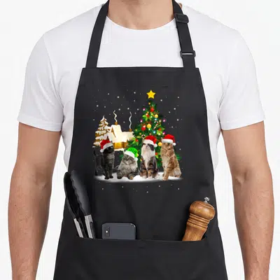 Tablier de cuisine noir avec quatre chats portant des bonnets de Noël devant un sapin illuminé.